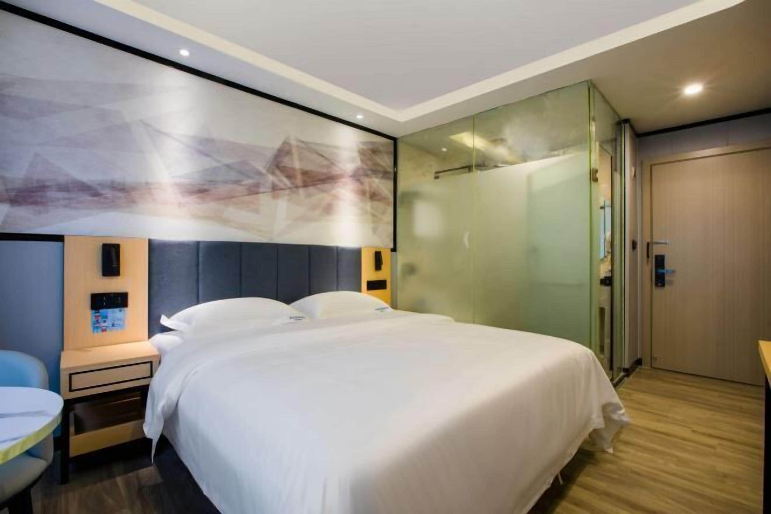 A modern hotel room with a large bed, a desk, and a glass partition wall.