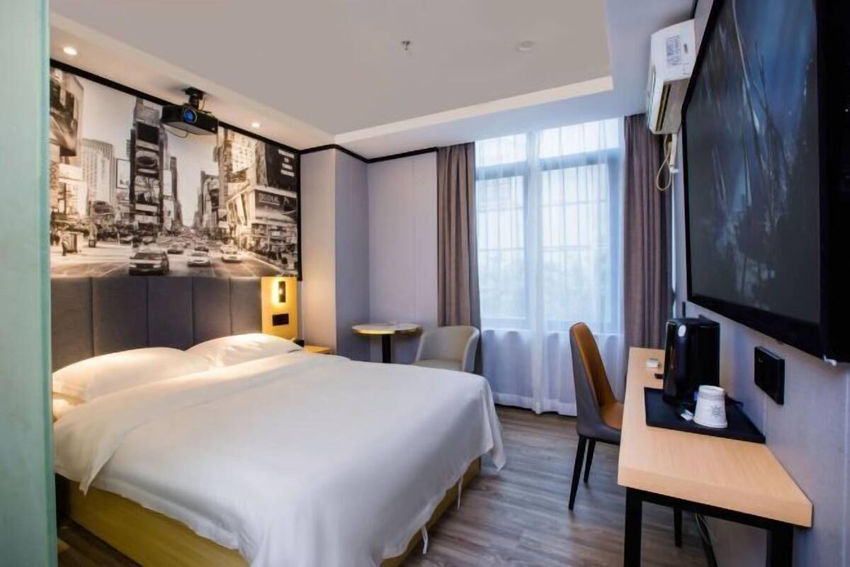 A hotel room with a bed, a desk with a computer, a TV, and a window with curtains.
