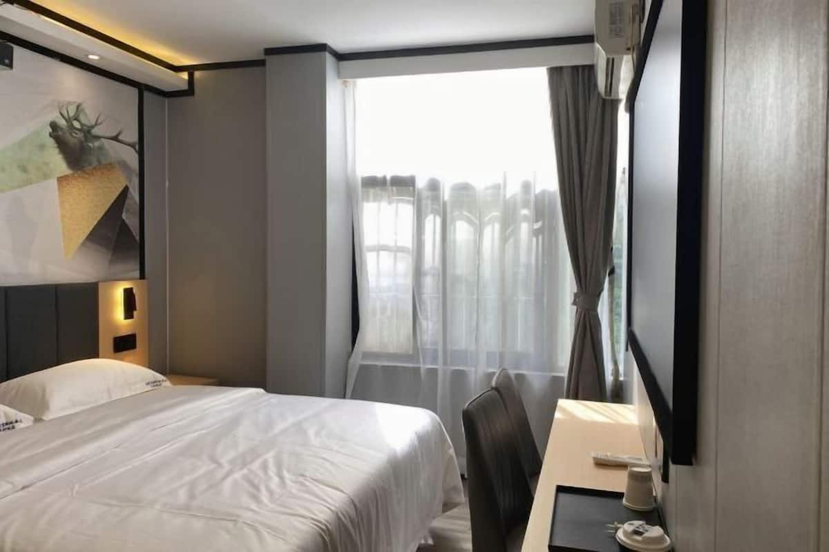 A hotel room with a bed, a desk, a chair, a window with curtains, and a wall-mounted air conditioning unit.
