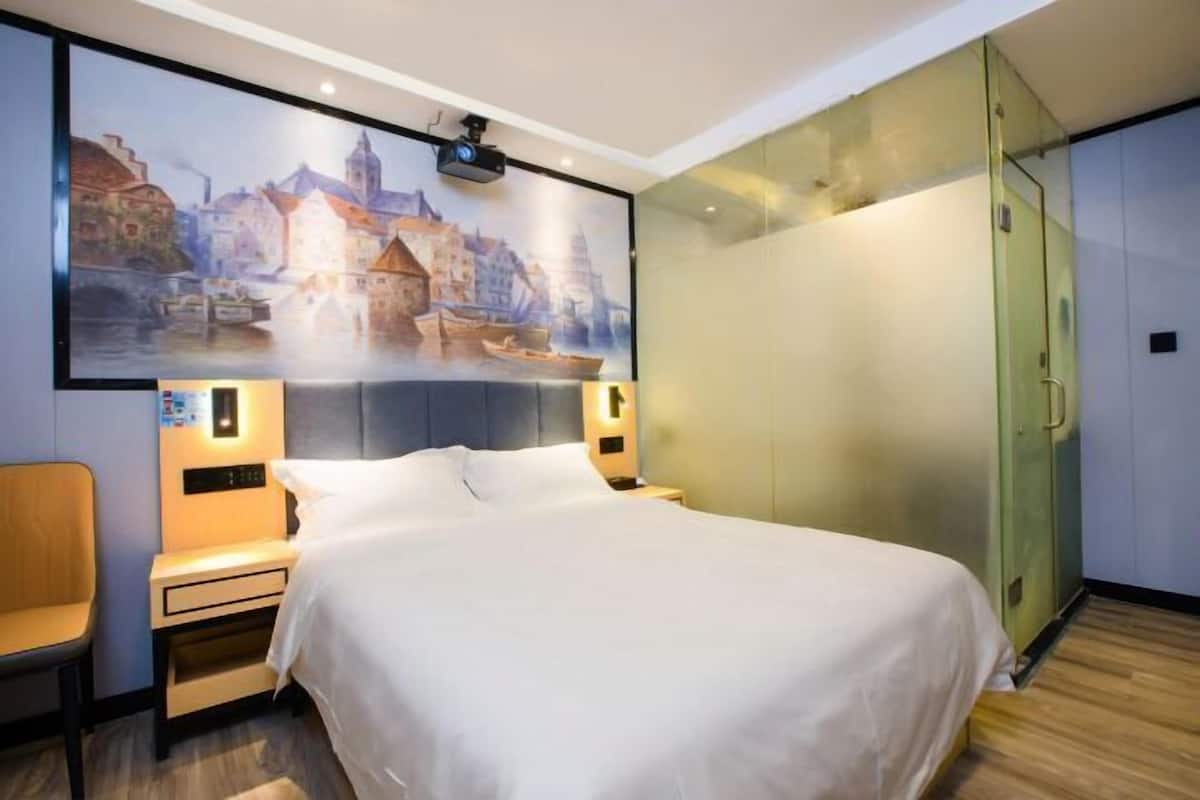 A hotel room with a bed, a chair, a nightstand, a wall-mounted projector, and a sliding glass door closet.
