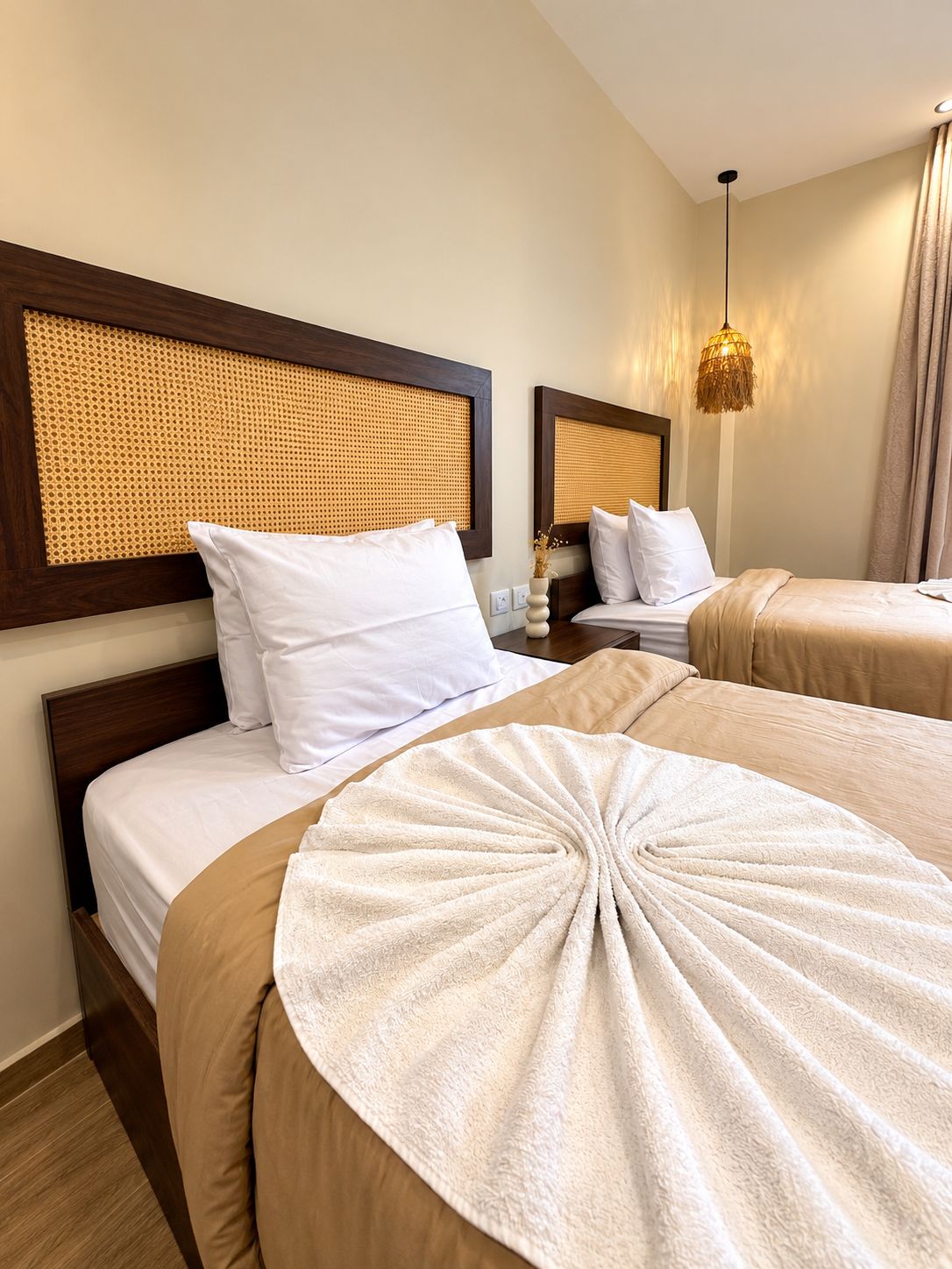 A hotel room with two beds, a rattan headboard, a bedside lamp, and a decorative towel.