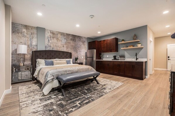 A modern bedroom with a large bed, bedside tables, and a bench. Adjacent is a kitchen with dark wooden cabinets, a refrigerator, and a countertop.