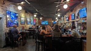 Zona de bar con varios televisores, paredes de ladrillo y clientes sentados en mesas.