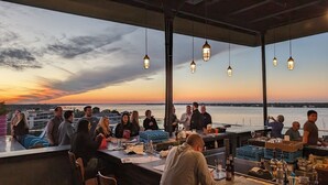 Eine Dachterrasse mit Blick auf den Sonnenuntergang über dem Wasser, Tische zum Essen gedeckt und hängende Pendelleuchten.