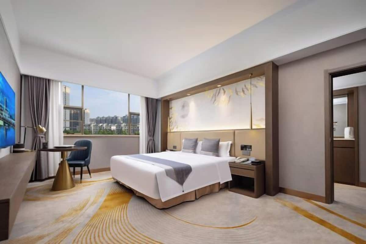 A hotel room with a large bed, a desk, a chair, a nightstand, and a view of the city.