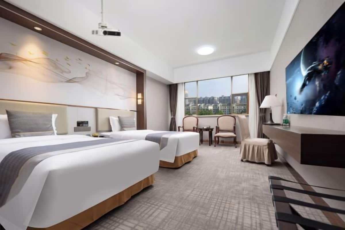 A hotel room with two beds, a large window with a city view, a desk with a lamp, and a painting on the wall.