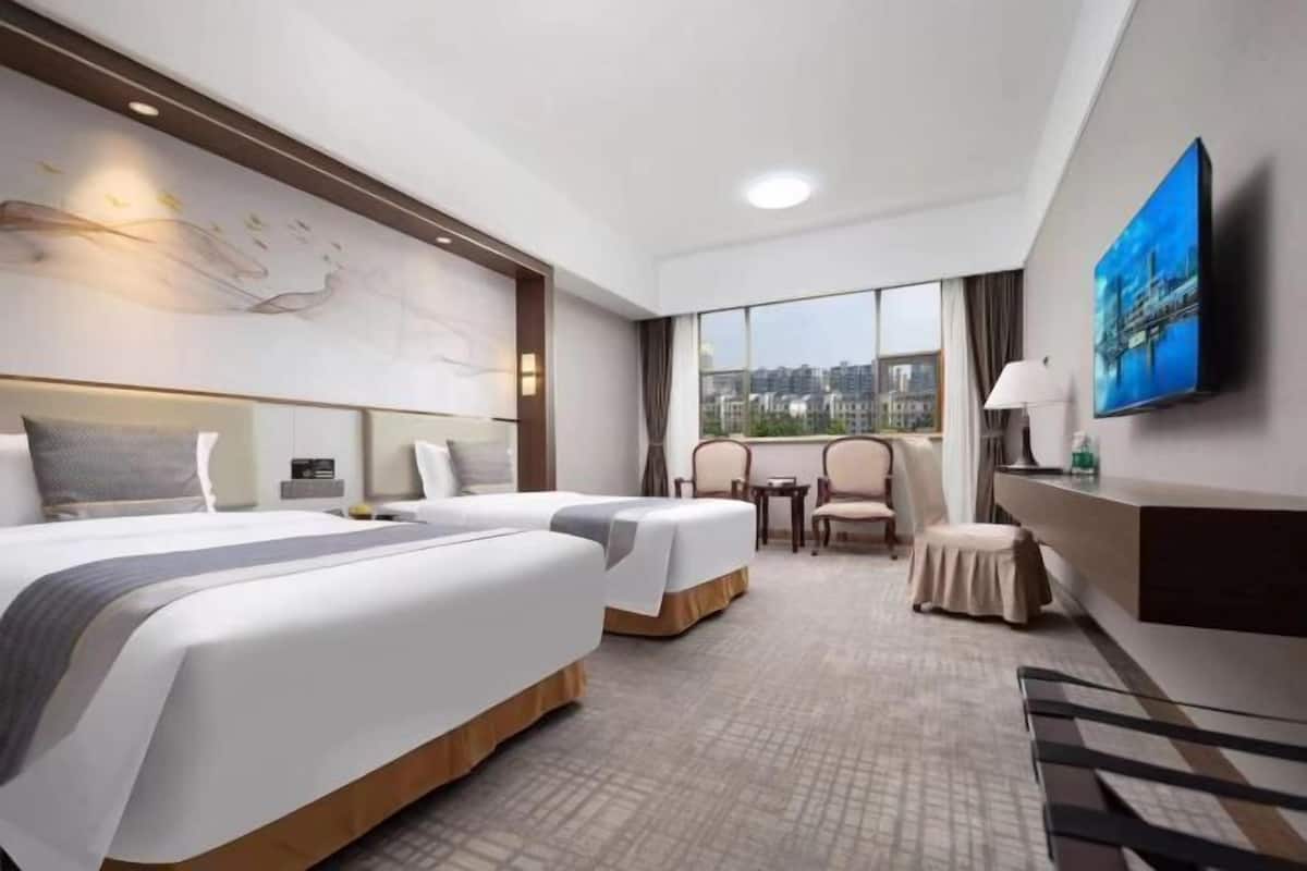 A hotel room with two beds, a desk, a chair, and a large window offering a city view.