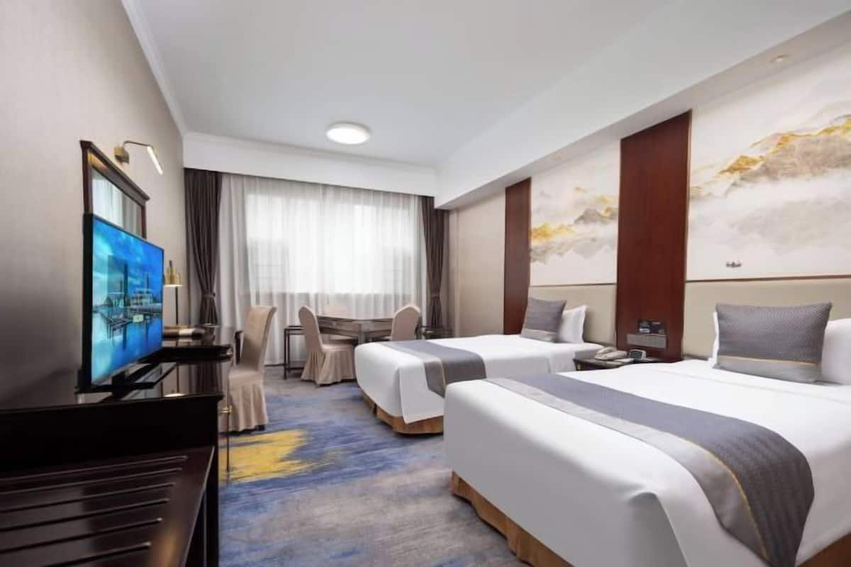A hotel room with two beds, a TV, a desk, and a large window with curtains.