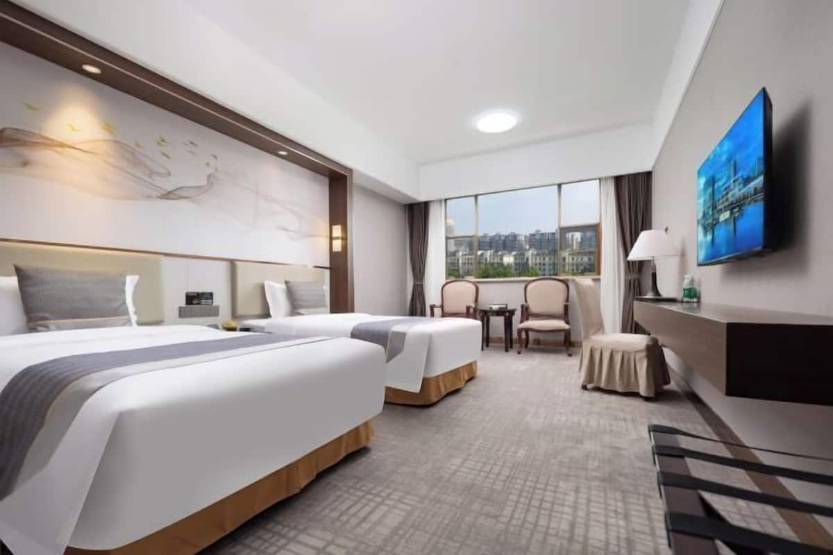 A hotel room with two beds, a desk, a chair, a small table, and a large window offering a city view.