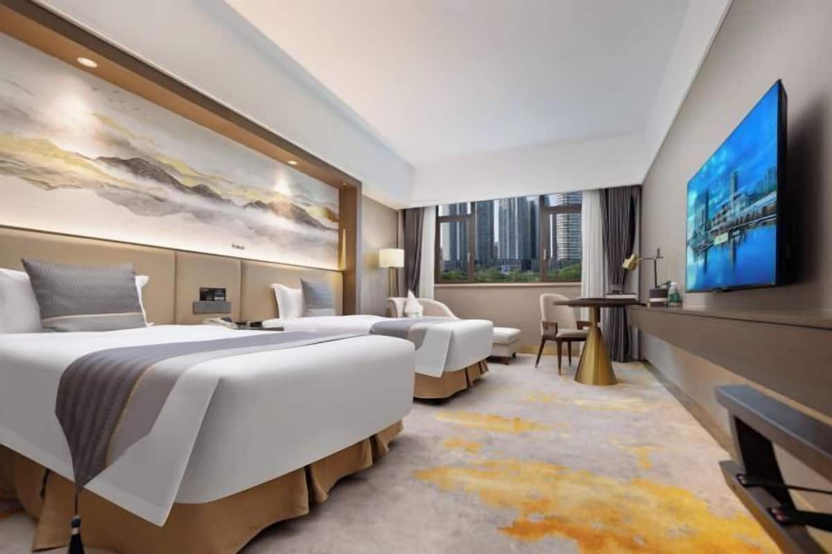A hotel room with two beds, a TV, a desk, and a large window with a city view.