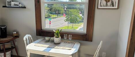 A dining area with a table and chairs, a potted plant, a wine glass, and a view of a street and greenery outside.
