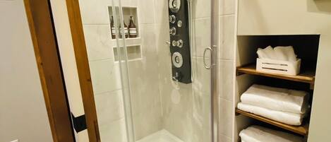 A modern bathroom with a glass-enclosed shower, white tiled walls, a wooden shelf with towels, and a small basket.
