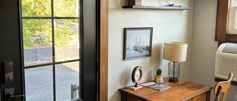 Eine schwarze Tür mit Fußmatte, ein Holztisch mit Lampe, ein Stuhl, ein an der Wand montierter Fernseher und ein kleiner Blumentopf.