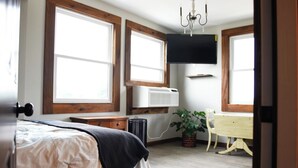A bedroom with a bed, large windows, an air conditioning unit, a TV, a plant, and a wooden chair.