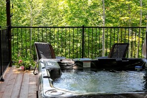 A hot tub with two chairs on a wooden deck, surrounded by a metal fence and lush greenery. - Pur Nature Eastman et Spa (Eastman)