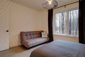 A bedroom with a bed, a sofa, a window with a view of trees, and a hanging light fixture. - Pur Nature Eastman et Spa (Eastman)