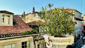 Un balcón con una urna blanca grande y una planta, con vistas a un paisaje urbano con edificios históricos y un cielo azul despejado.