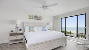 Ein Schlafzimmer mit einem großen Bett, einem Nachttisch, einer Lampe, einem Deckenventilator, einem Schaukelstuhl und Blick auf den Strand.