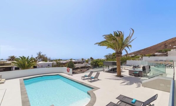 Piscina no terraço com espreguiçadeiras, uma palmeira e vista para o oceano e as colinas.