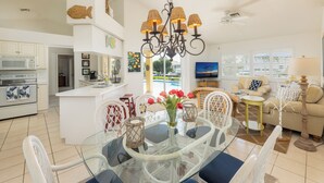 A modern kitchen with white cabinets, a dining area with glass tables and chairs, a living room with a sofa and a TV, and a view of the outdoors through large windows.