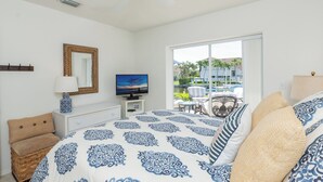 A bedroom with a bed, a television, a lamp, a chair, and a view of a pool and buildings outside.