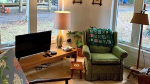 A living room with a television, a green armchair, a wooden coffee table, and a lamp. There are two windows with a view of trees and a parked vehicle outside.