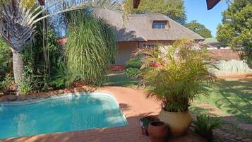 Relax in a serene backyard setting surrounded by lush greenery and a well-maintained lawn, featuring a swimming pool and a rustic house with a thatched roof.