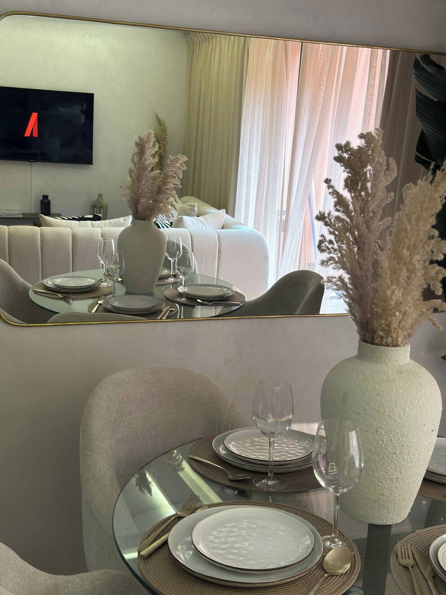 A dining area with a modern table set for two, a vase with dried plants, and a large mirror reflecting the room.