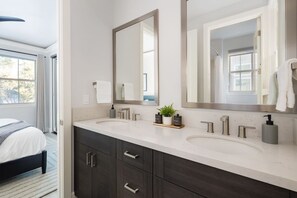 A modern bathroom with two sinks, a large mirror, and a bed with a view to the outdoors.