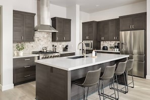 A modern kitchen with dark cabinetry, a central island with bar stools, a stainless steel range, and a large refrigerator.