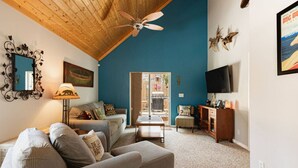 A living room with a ceiling fan, a sofa, a coffee table, a TV, and a door leading to an outdoor area. - Blissful Bay Cabin + Block from Boulder Bay (Big Bear Lake)