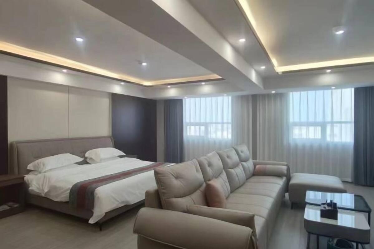 A modern hotel room with a bed, a sofa, a coffee table, and large windows with curtains.