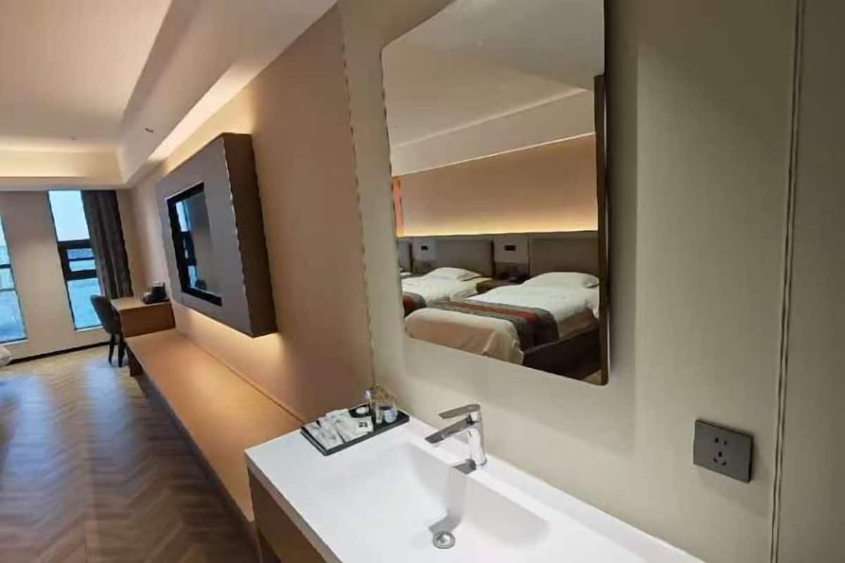 A modern hotel bathroom with a large mirror, a sink, and a bed visible in the reflection.
