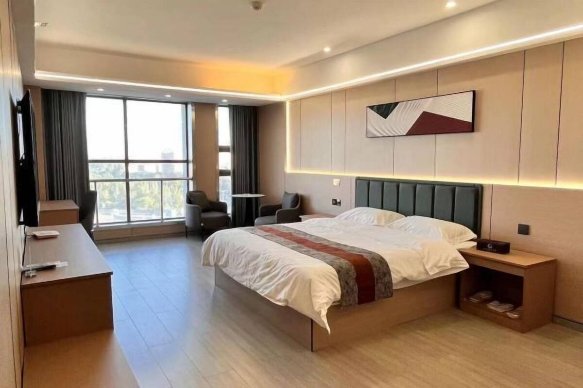 A modern hotel room with a large bed, a desk, a chair, and a view of the city through large windows.
