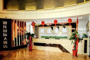 Zona de recepción de un hotel con un mostrador, plantas en macetas y un cartel grande con caracteres chinos.