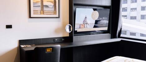 Una habitación de hotel moderna con un televisor de pantalla plana grande montado en la pared, un escritorio con una silla, y un cuadro enmarcado sobre el escritorio.
