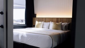 A neatly made bed with white linens and multiple pillows, a wooden headboard, and a window with a view of buildings. - One-Bedroom Apartment in CBD (Newcastle)