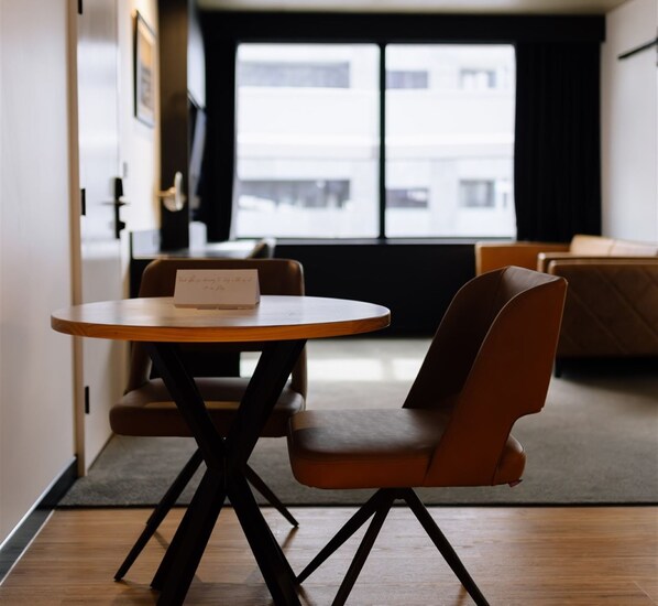 A compact living space with a round wooden table, two brown chairs, a small rug, and a view of buildings through a window.