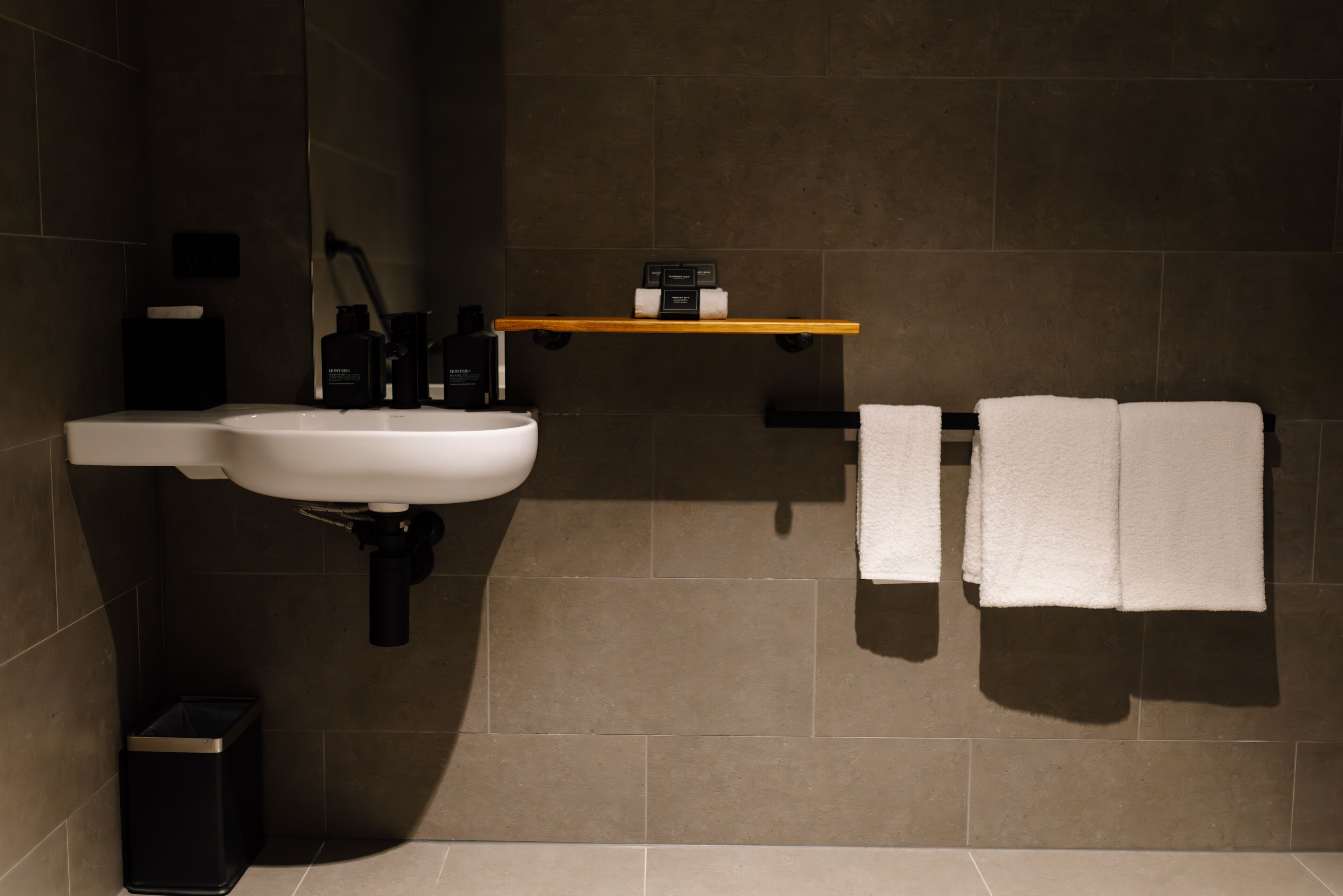 A bathroom with a white sink, black trash bin, and a wooden shelf with toiletries.