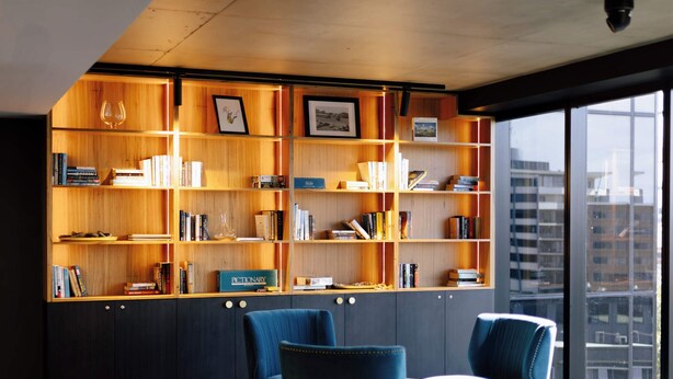 A modern reading area with a large bookshelf, two blue armchairs, and a city view through large windows. - One Bedroom Accessible In CBD (Newcastle)