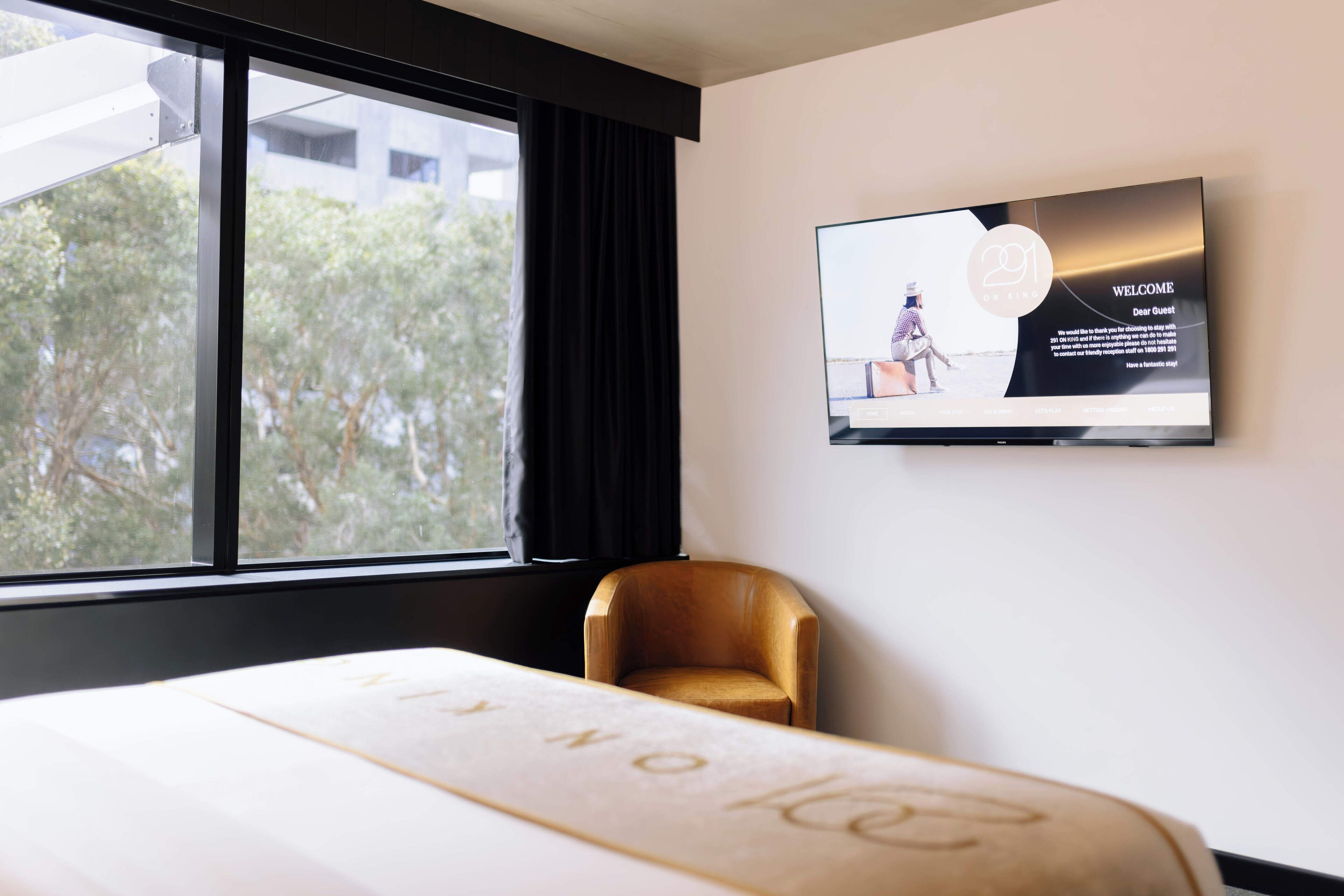Une chambre d’hôtel dotée d’une grande fenêtre, d’un téléviseur à écran plat fixé au mur, d’une chaise en bois et d’un lit avec une tête de lit.