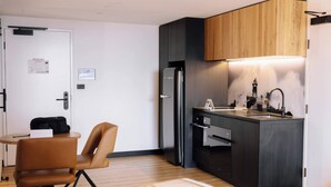 A compact kitchen with a modern black refrigerator, wooden cabinets, and a small dining area with a brown chair and a table. - Two Bedroom Interconnect In CBD (Newcastle)