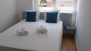 Una cama bien hecha con sábanas blancas y toallas enrolladas, una mesita de noche con lámpara, y un papel tapiz estampado.