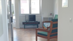 Amplia sala de estar con suelo de parqué tipo espiga, televisor, una mesita y una ventana con persianas.