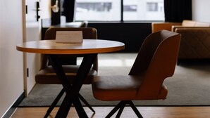 A modern room with a round wooden table, two brown chairs, and a view of buildings through a large window. - Two Bedroom Interconnect Deluxe In CBD (Newcastle)