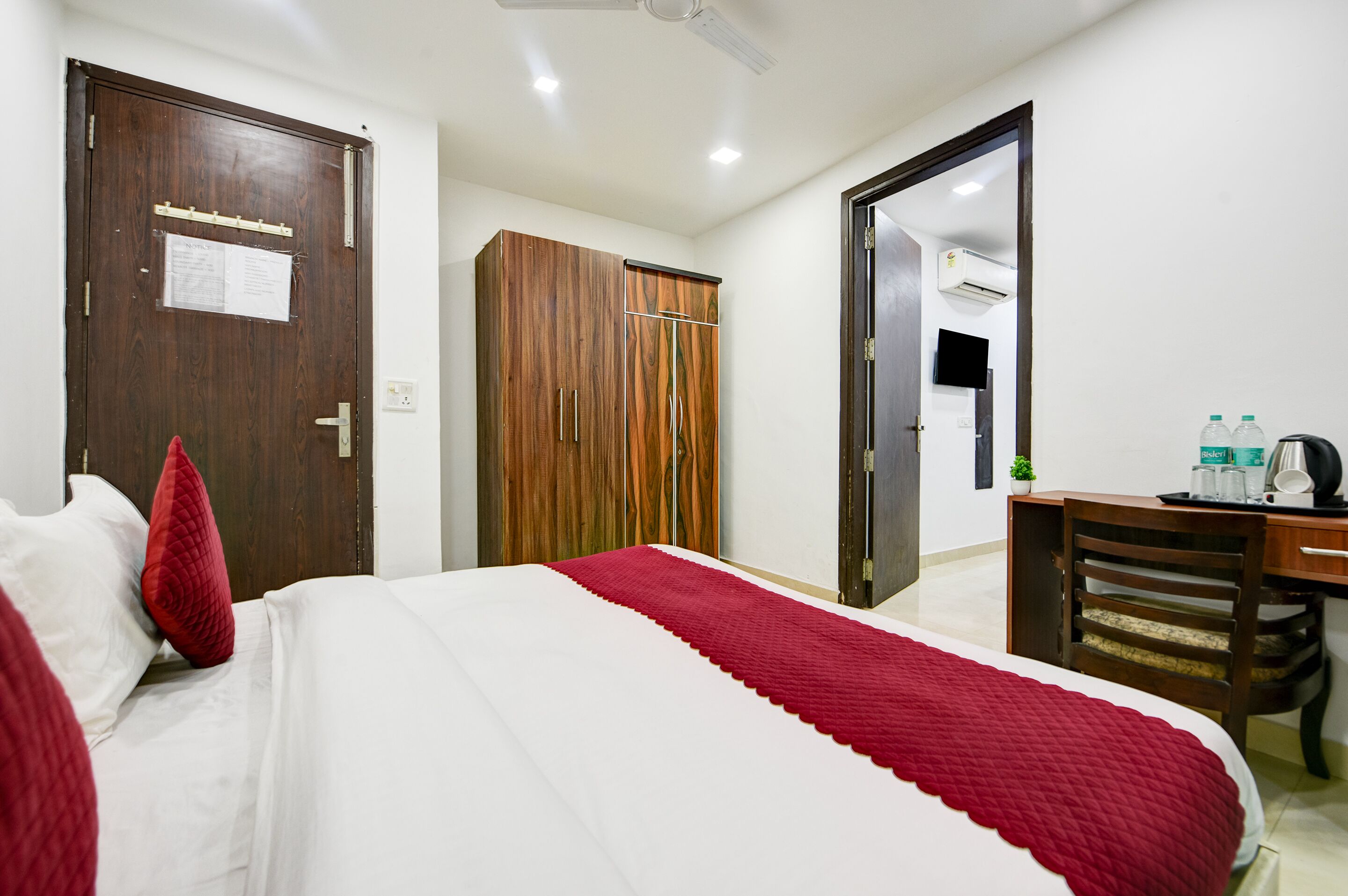 A hotel room with a bed, a wardrobe, a desk with a chair, and a small table with bottles and a kettle.