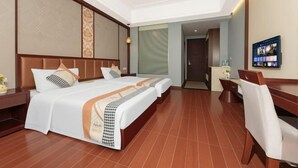 Quarto de hotel com uma cama grande, uma escrivaninha e uma cadeira. Há uma televisão na parede e uma janela com cortinas.