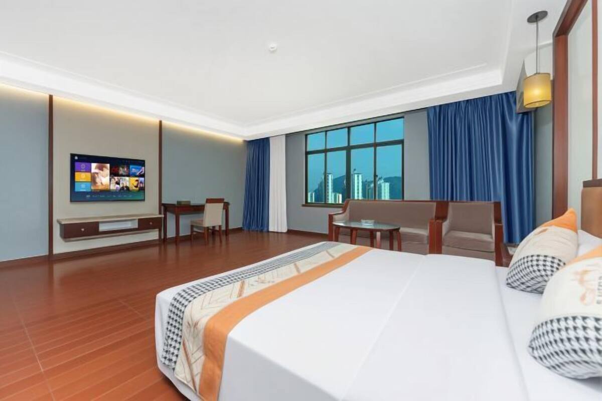 A hotel room with a large bed, a desk, a chair, a TV mounted on the wall, and a window with a city view.