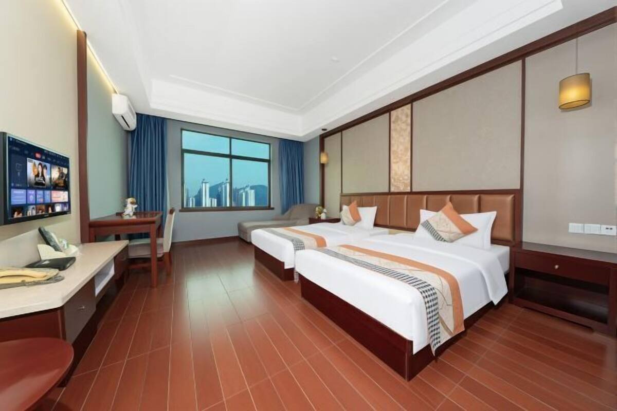 A hotel room with two beds, a desk, a chair, a TV, and a window with a city view.