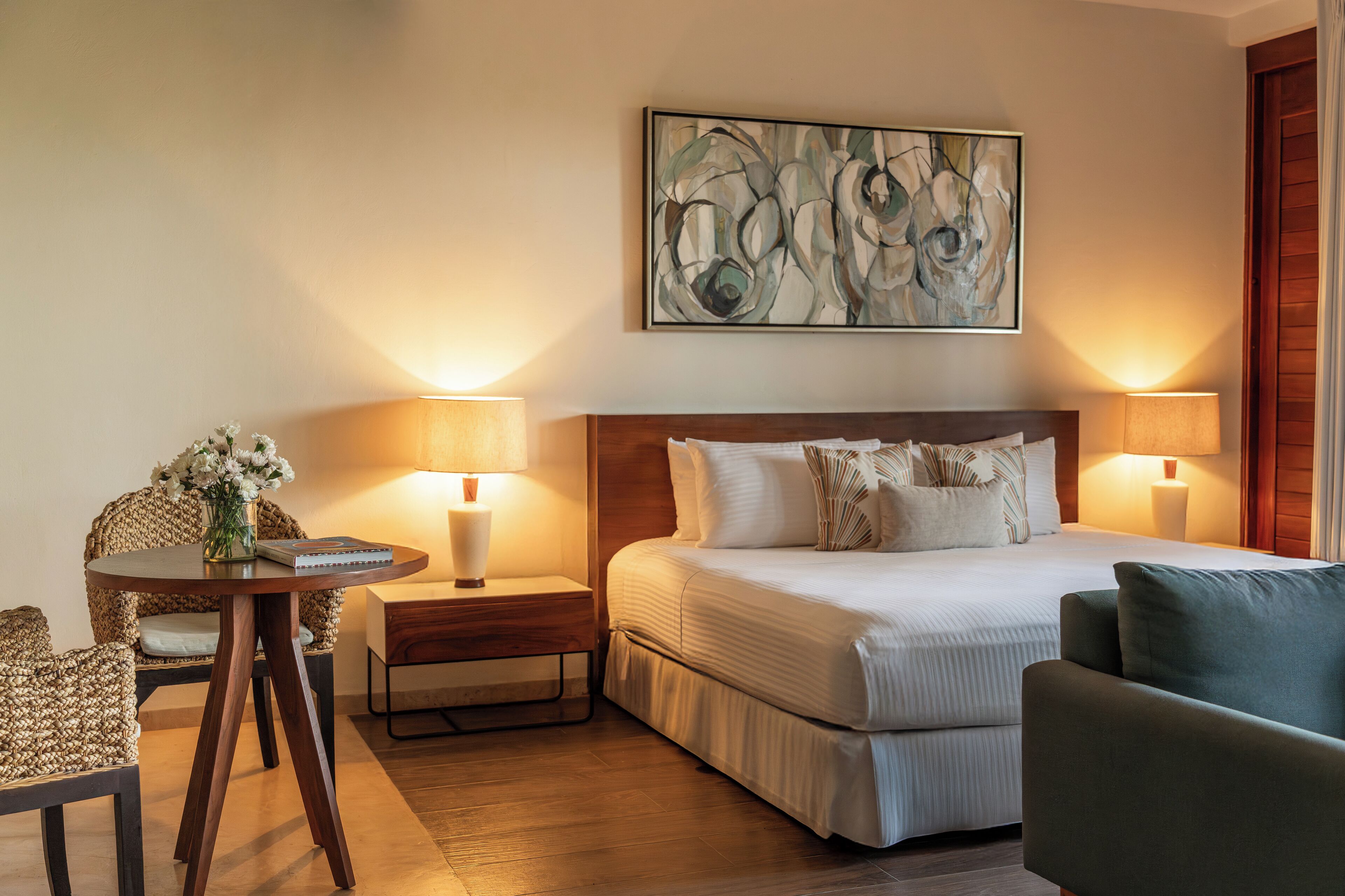 Una habitación de hotel con una cama, dos lámparas de noche, una mesa redonda con lámpara, una silla y un cuadro abstracto colgado en la pared.